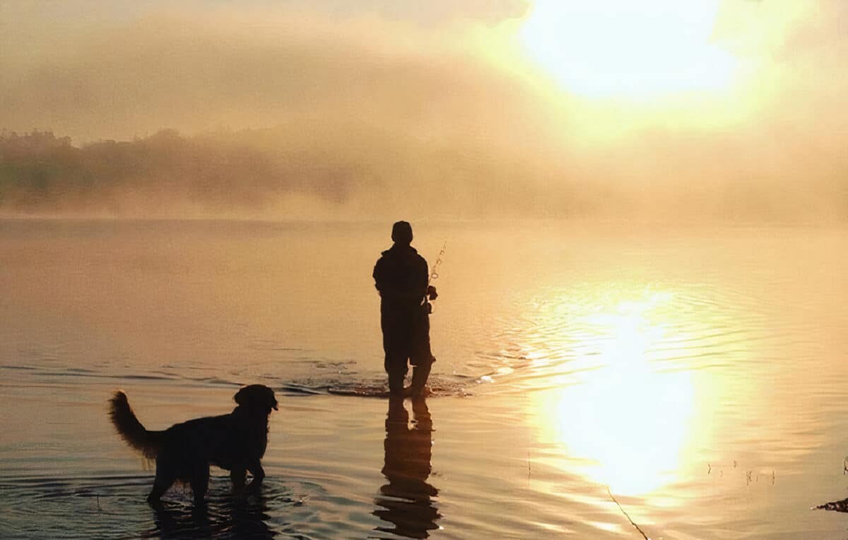 Rybář se brodí vodou při západu slunce se psem