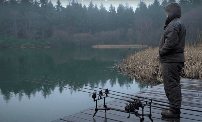 Rybář stojí u nahozených prutů v teplém obleku Avid Carp