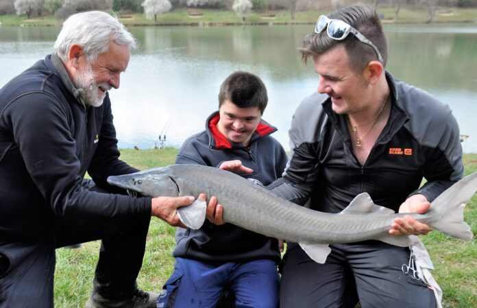 Rybáři ukazují úlovek jesetera handicapovanému rybářovi