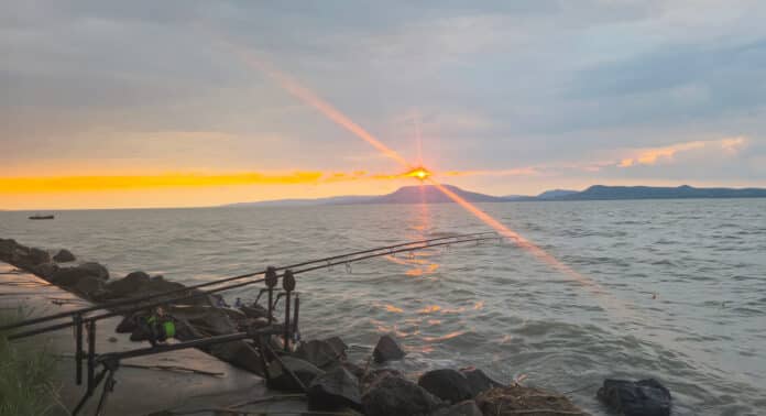 Sunset over a calm lake with fishing rods propped along a rocky shore.