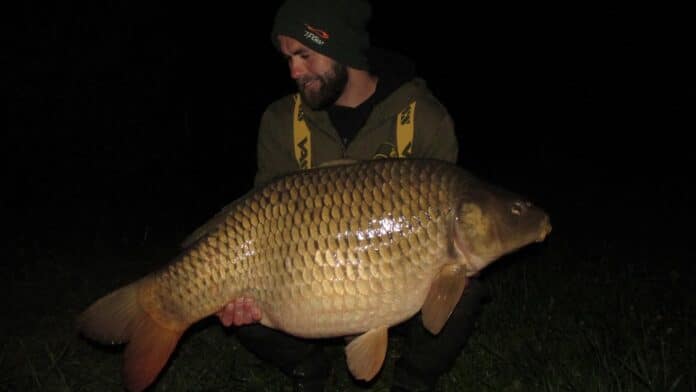 Man in a dark beanie and olive jacket holding a large golden carp at night, wet and glistening from water.