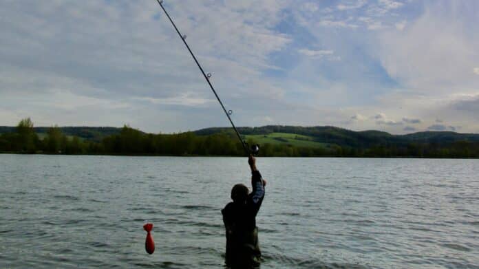Person standing in knee-deep water, reeling in a long fishing rod over a lake with distant hills and a red bobber floating nearby