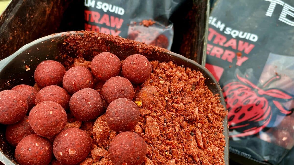 Close-up of red fishing bait boilies (spherical bait) with a red powder coating in a bowl, some crumbs spilled nearby.