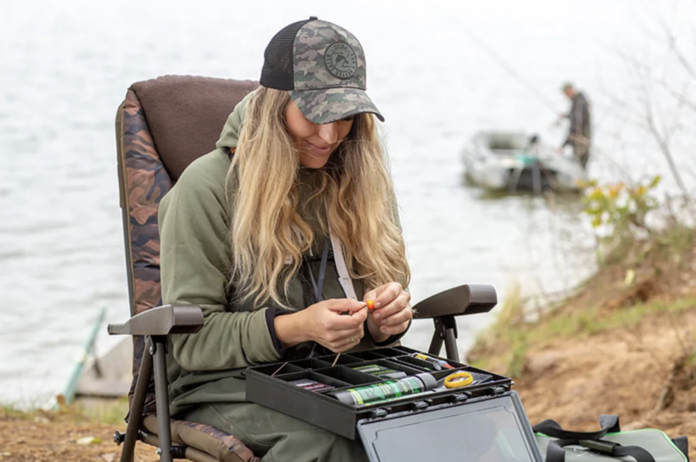 Woman in a camo cap and green hoodie sits in a folding chair by a lake, assembling fishing gear from a tackle box.