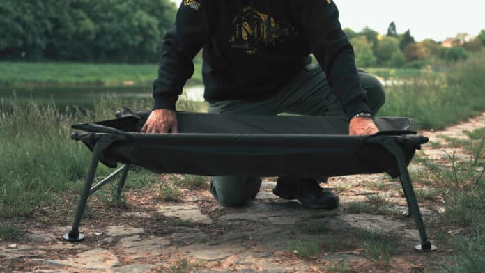 Person kneeling on the ground and setting up a portable camping cot in a grassy outdoor area near a body of water.
