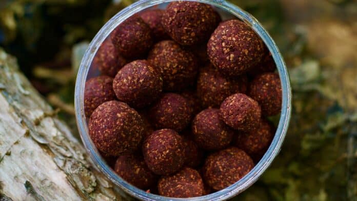 Jar of reddish-brown energy balls coated with nut fragments, resting on a rustic surface