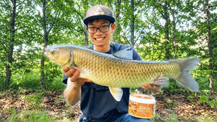 Smiling man with glasses holds a large golden fish horizontally outdoors, trees in the background.