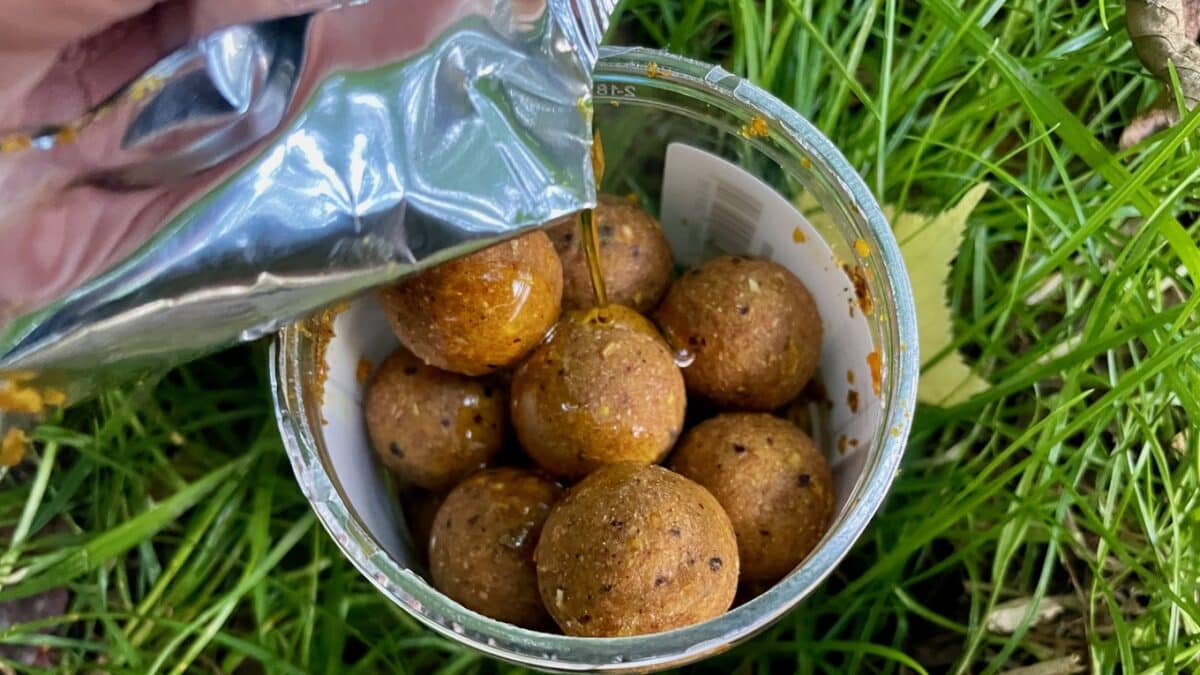 Open jar of brown boilie-style fishing bait balls on grass, with a foil pouch pouring orange liquid over them.