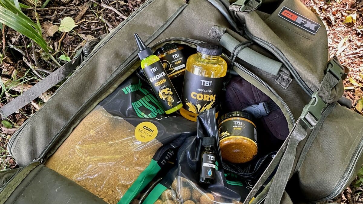 Open green tackle bag exposing fishing bait jars, corn flavour boosters, and corn mix packets on a forest floor.