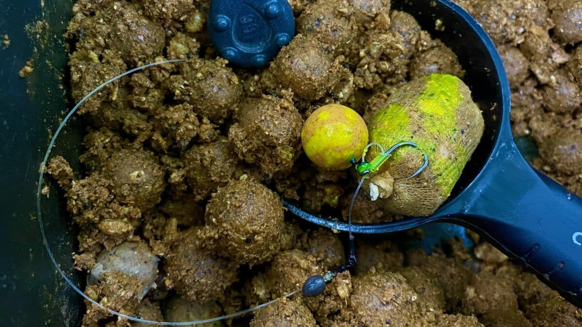 Close-up of fishing rig in a muddy scoop: yellow float, green hook bead, and two hooks attached to a wire leader amidst brown bait balls.
