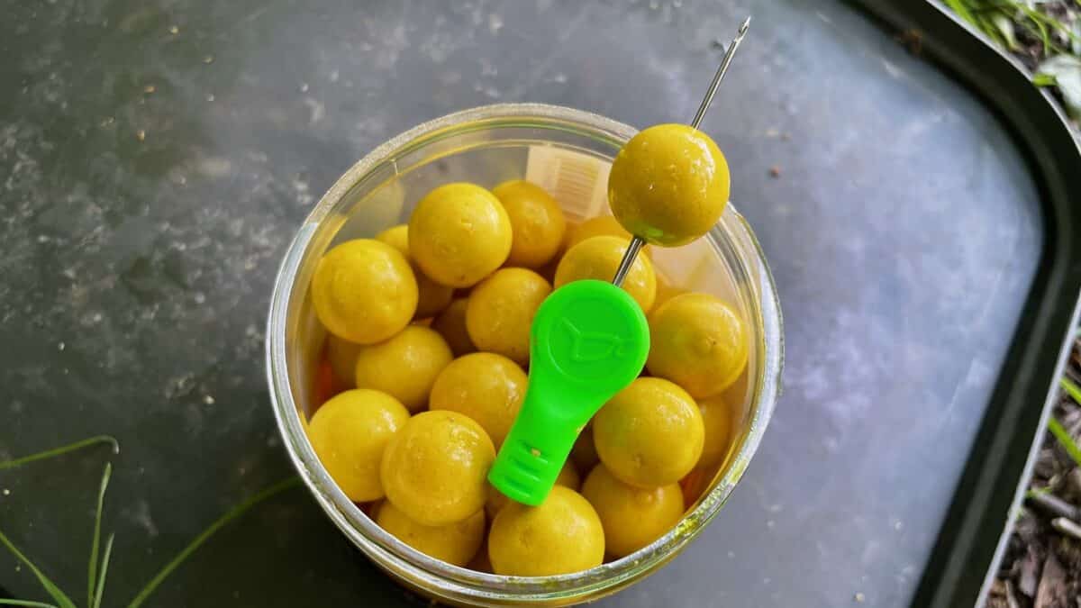 Open jar of yellow-green olives with a metal skewer bearing an olive and a bright green plastic scoop inside.