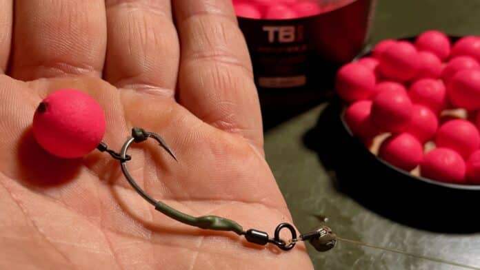 Close-up of a bright pink fishing bead on wire being held in a hand, with a bowl of pink beads in the background.