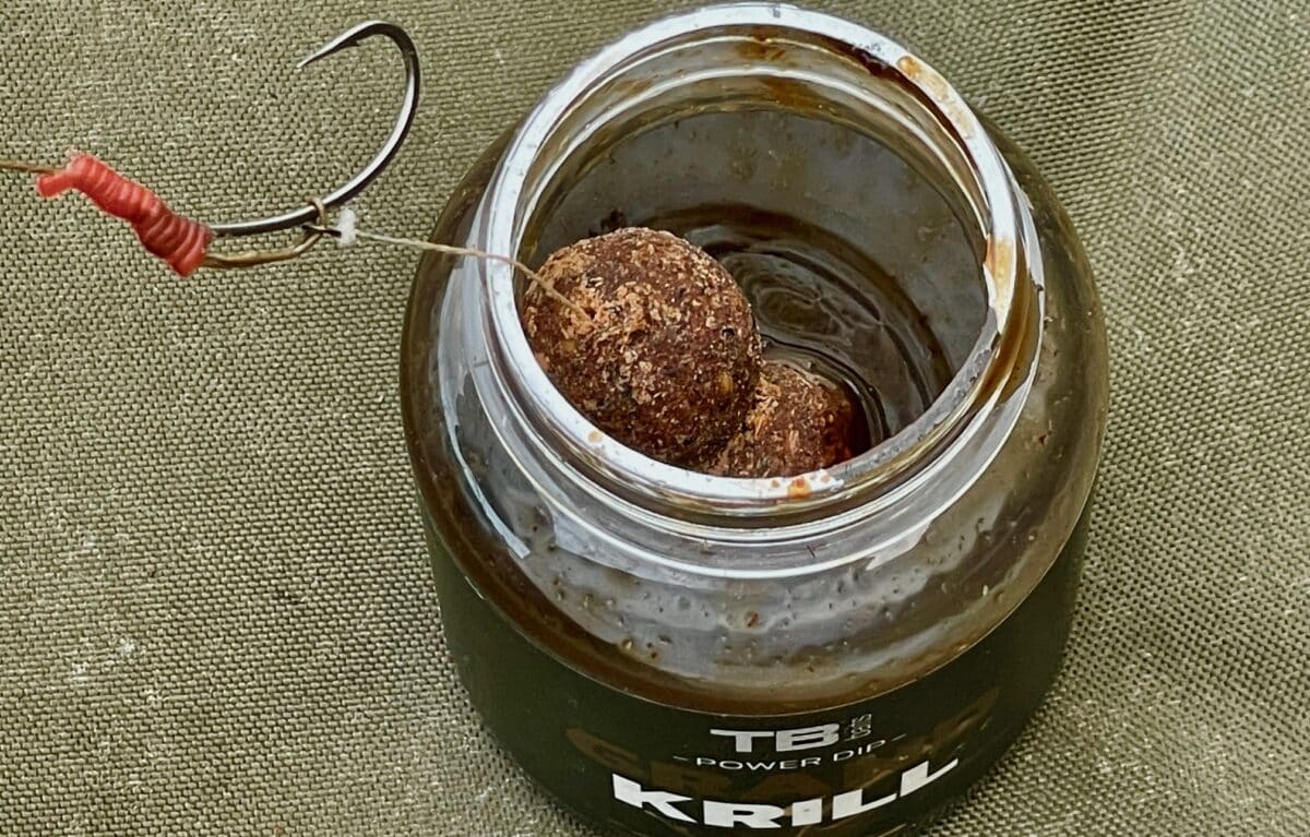 Close-up of a glass jar with a metal lid, containing a brown textured fishing lure attached to a line wrapped in red thread, set on a textured fabric surface.