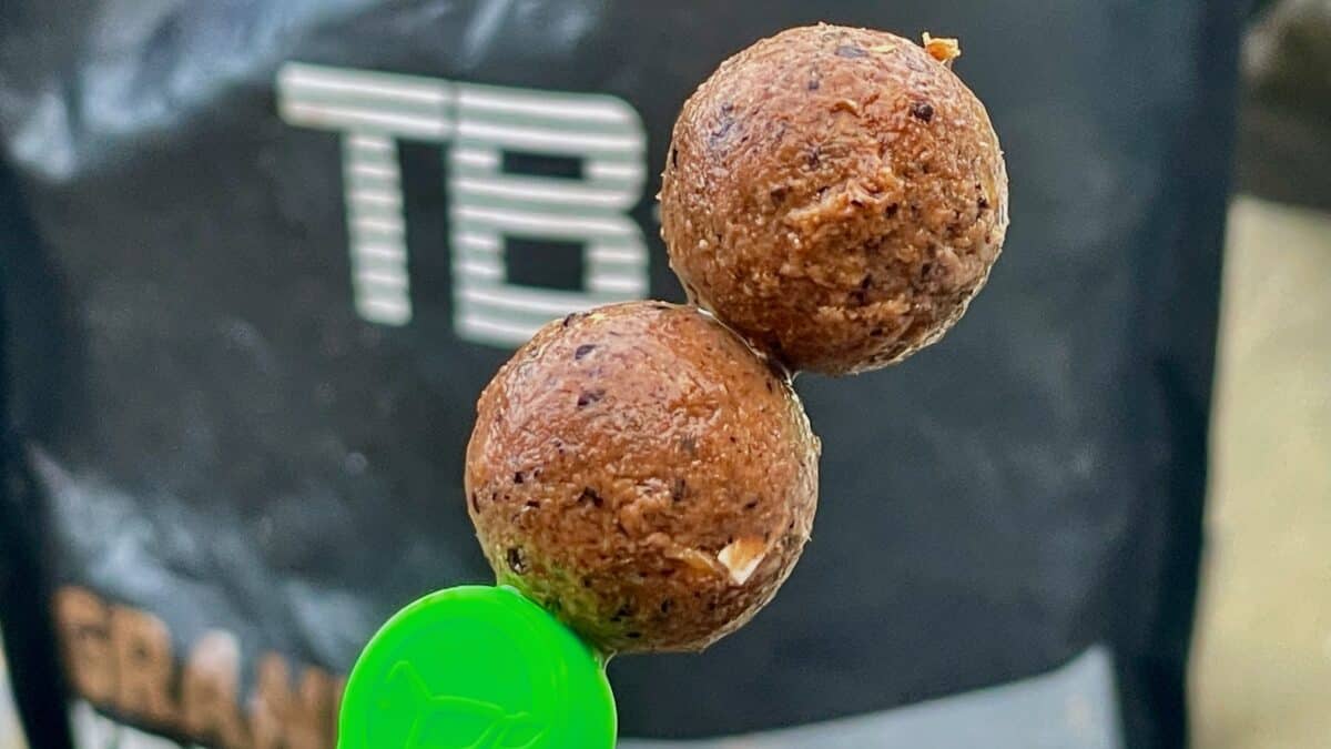 Three brown fried meatballs on a skewer, held up against a dark background with a green lid visible in the foreground.