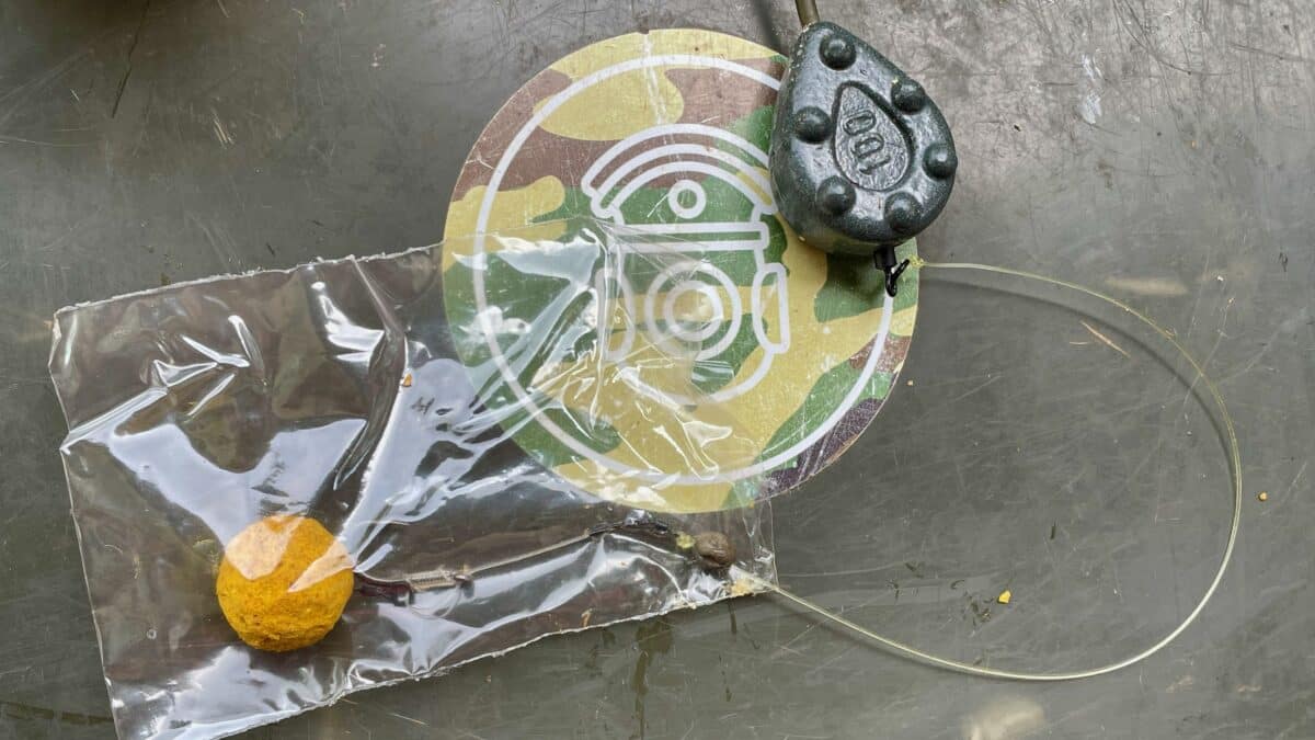 Close-up of a multicolor circular sticker on a scratched metal surface, with a gray studded fishing lure resting on it and a plastic bag containing an orange ball nearby.