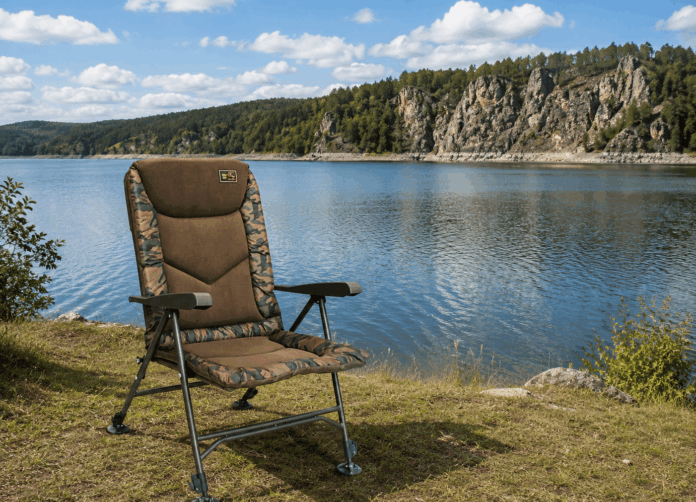 Camouflage padded lawn chair on a grassy lakeshore, with calm water and rocky cliff backdrop.
