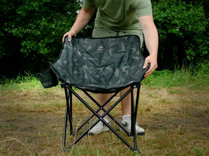 Person unfolding a camouflage folding camping chair outdoors on a grassy area, hands adjusting the fabric.