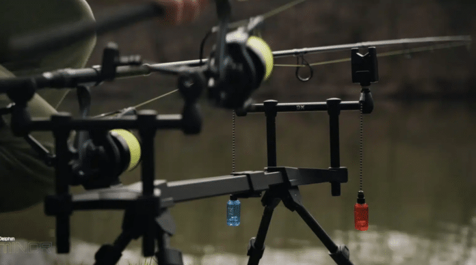 Close-up of a fishing rod rig with reels, lines, and two hanging colored lure capsules (blue and red) near a calm body of water