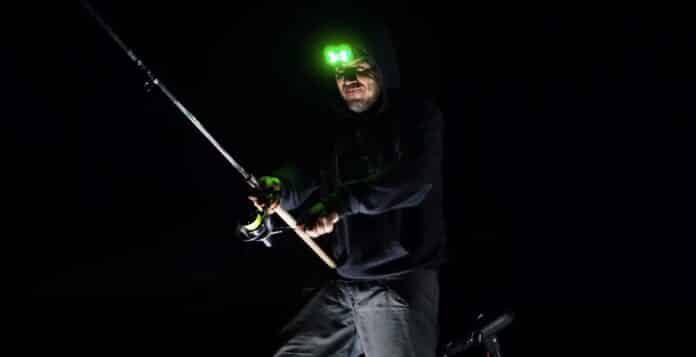 Man fishing at night wearing a headlamp that glows green, holding a fishing rod in a dark setting