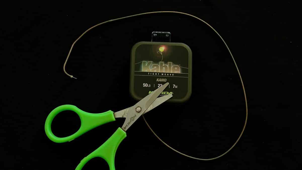 Green-handled scissors cutting a thin fishing line, with a small 'Kable' line spool in the background on a dark surface.