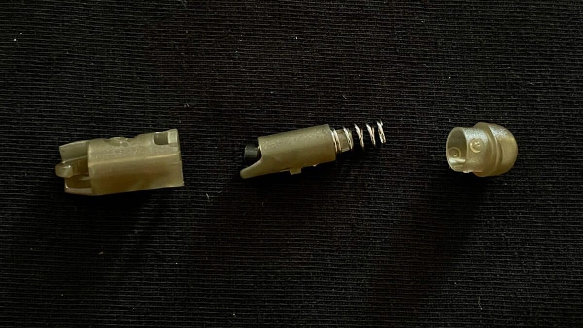 Three beige plastic connector parts with a metal compression spring laid out on a dark fabric surface, showing an exploded small electrical component.