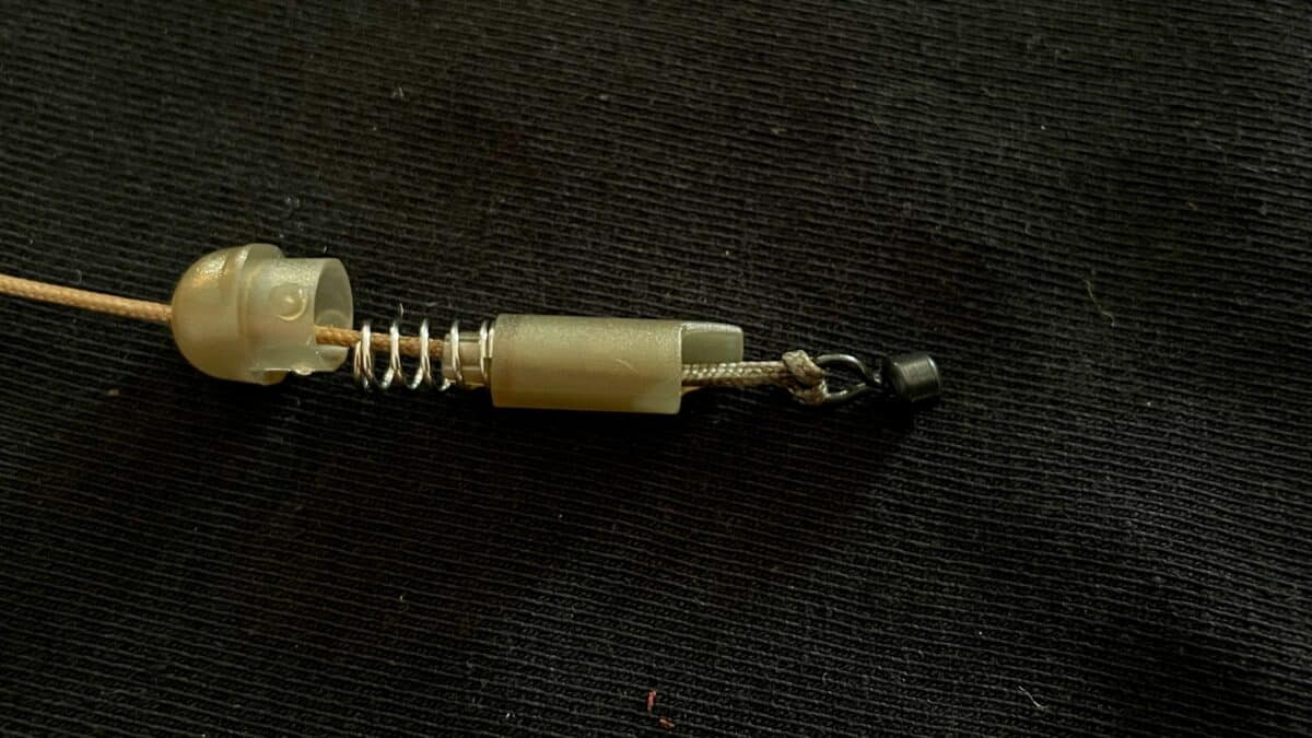 Close-up of a small beige plastic component with a metal spring on a brown cord on a dark fabric surface.