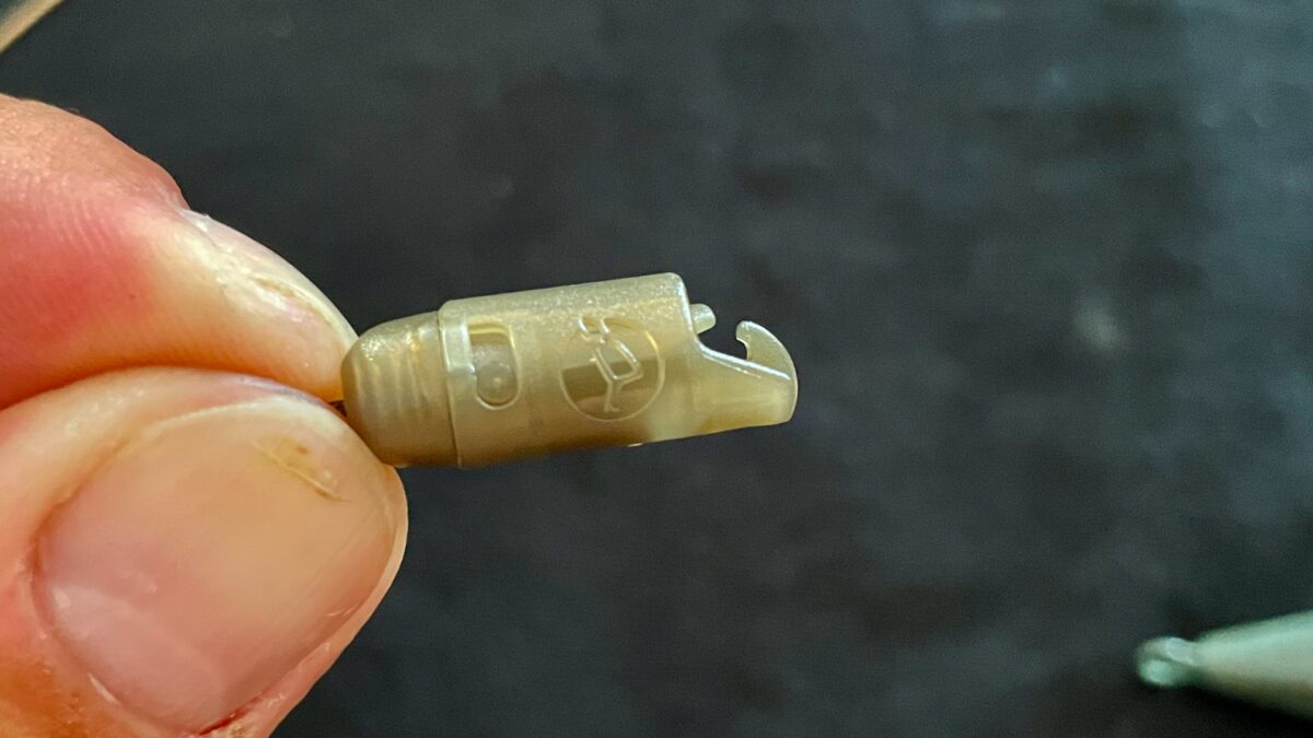 Close-up of a small beige plastic component held between fingers, showing a curved tip and a raised logo on the side against a dark background.