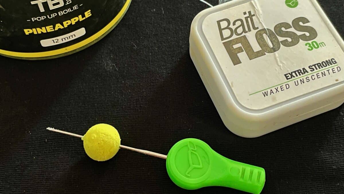 Close-up of fishing tackle: a small yellow boilie on a line, beside a bright green baiting needle and two bait tins labeled Pineapple pop-up and Bait Floss
