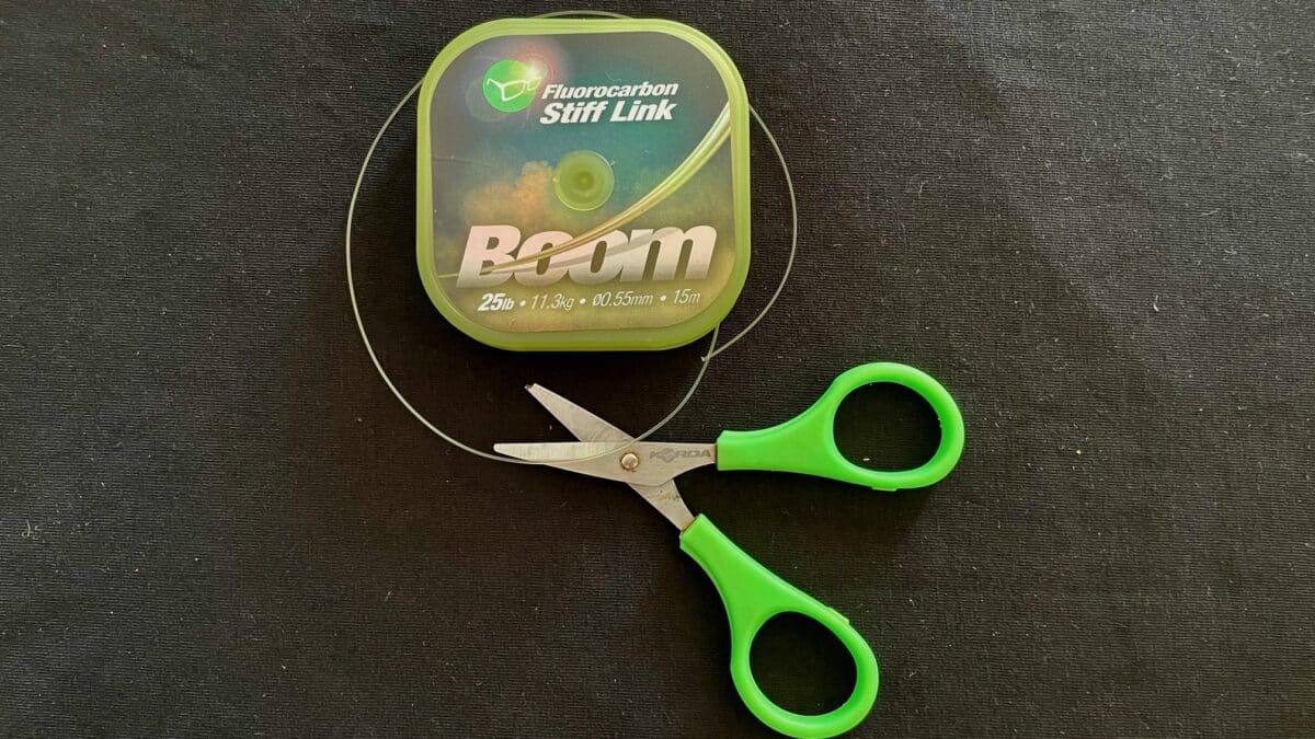 Green-handled scissors beside a neon-green case labeled Fluorocarbon Stiff Link fishing line on a dark textile surface.