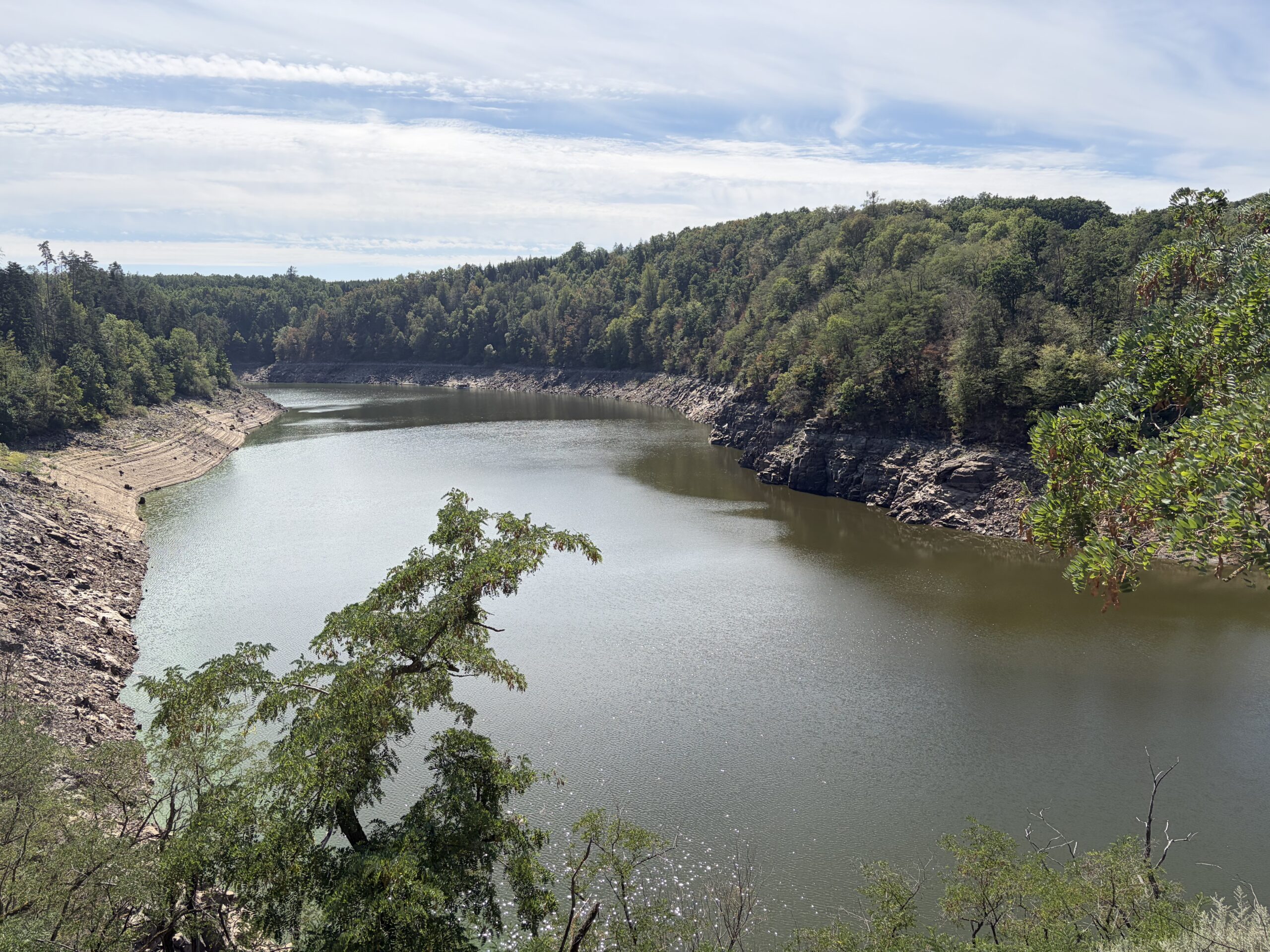 Wide river bends through rocky shoreline and forested hills under a blue sky with wispy clouds.