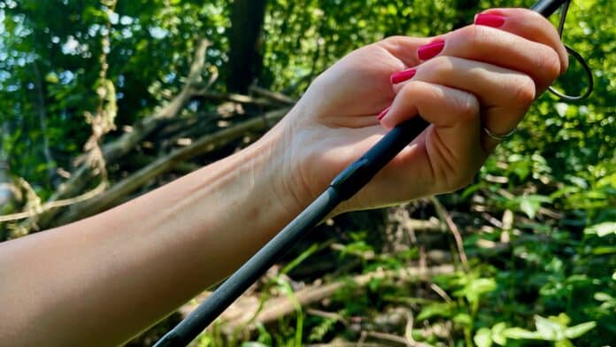 Close-up of a hand with pink nail polish gripping a fishing rod outdoors with green foliage in the background.