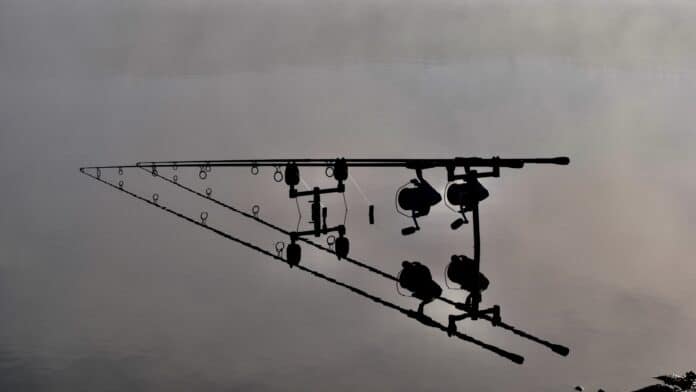 Silhouette of fishing rods and reels on a pier, reflected in calm water at dusk or dawn.
