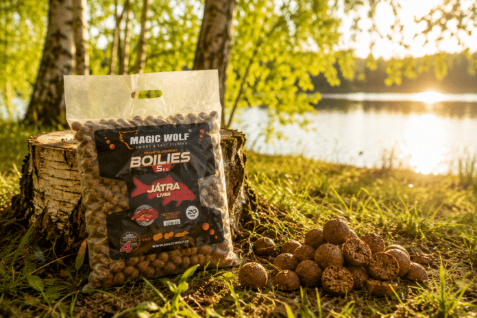 MAGIC WOLF Boilies bag on a tree stump by a sunlit lake, with a mound of boilies nearby.