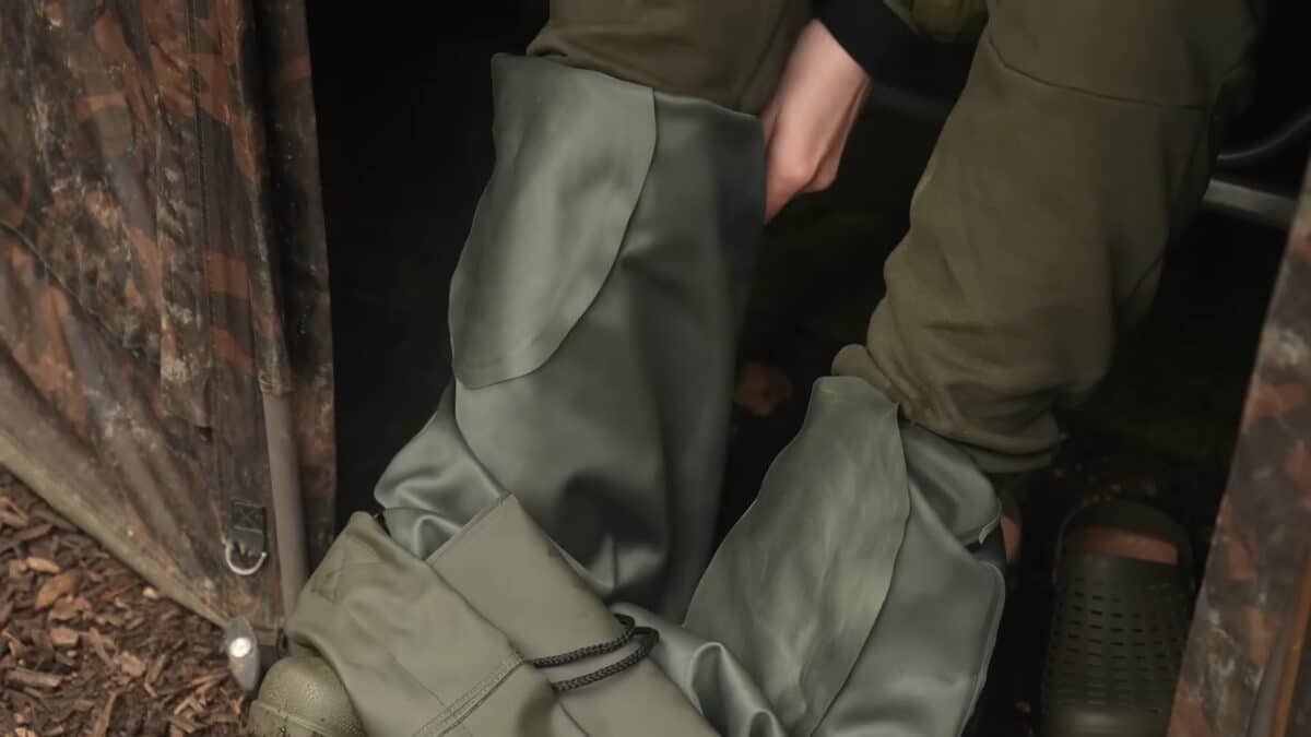 Person in olive-green pants and boots standing in a dark doorway with a green bag nearby.