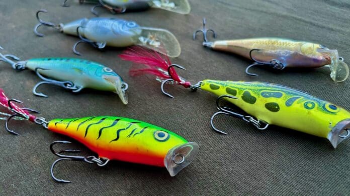 Assorted colorful fishing lures with treble hooks laid on dark fabric surface.