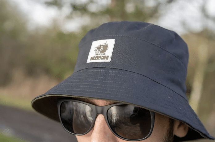 Close-up of a person wearing a navy bucket hat with a white patch reading 'NASH' and a dog logo, wearing black sunglasses outdoors.