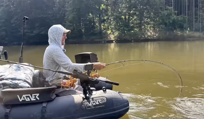 Person in a light hoodie and cap fishing from an inflatable boat on a calm lake, holding a long fishing rod with a bent tip as it reels in.