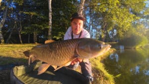 Man wearing a cap presents a large carp toward the camera with trees and a lake in the background