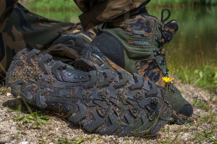 Camouflage hiking boot with muddy sole on a dirt path, near a small yellow flower in grass.