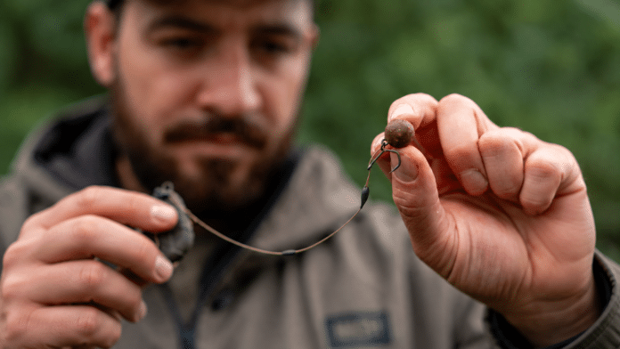 Man holding a fishing lure with a worm bait close to the camera, outdoors in a blurred green background.