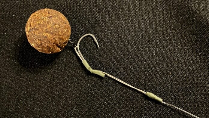 A round brown fishing weight or lure attached to a thin leader line with a small curved hook against a dark fabric background.