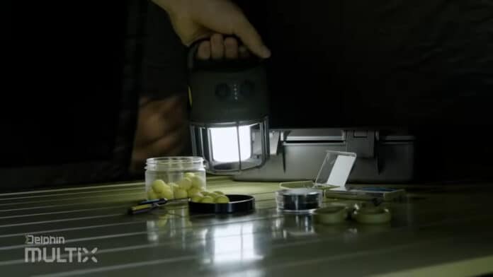 Person holding a lantern to illuminate fishing tackle laid out on a table, including bait balls in a jar and small gear, in a dark room.