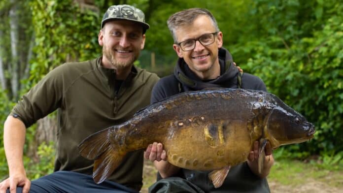 Two men outdoors smiling while holding a large carp, with a green, wooded background.