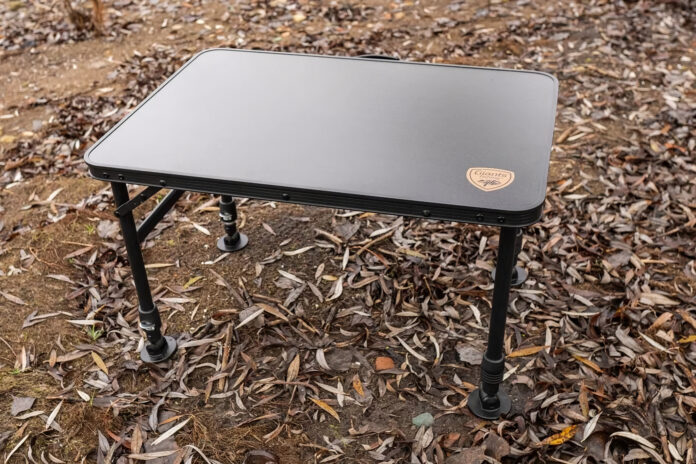 Outdoor folding camping table with black legs on a leaf-covered ground outdoors are visible in the image, the gray top catching light in a natural setting, suggesting a portable workspace.