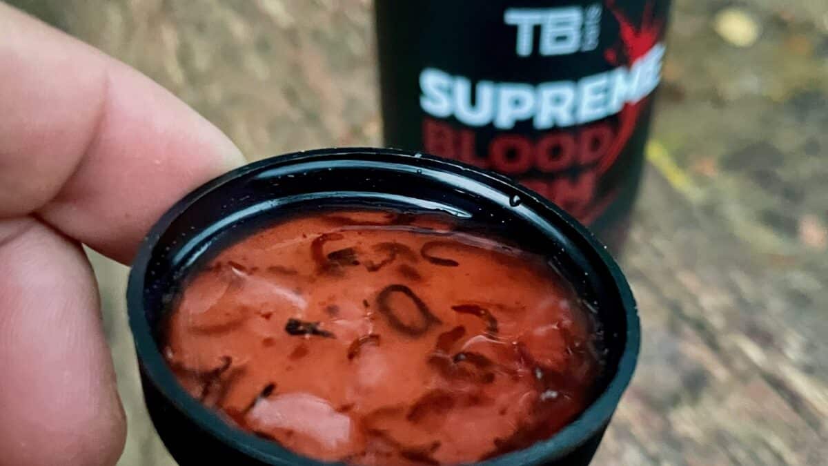 Close-up of a red glossy paste inside a black lid held by fingers, with a blurred bottle in the background.
