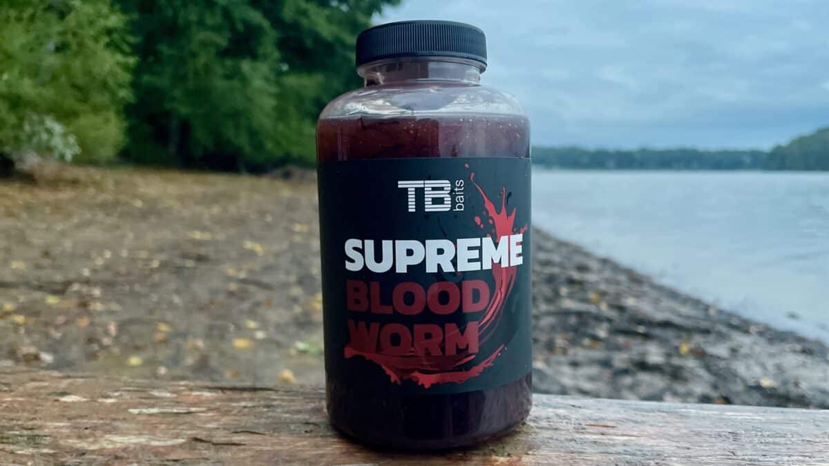 Close-up of a bottle labeled 'Supreme Blood Worm' on a weathered wooden surface by a lakeshore.