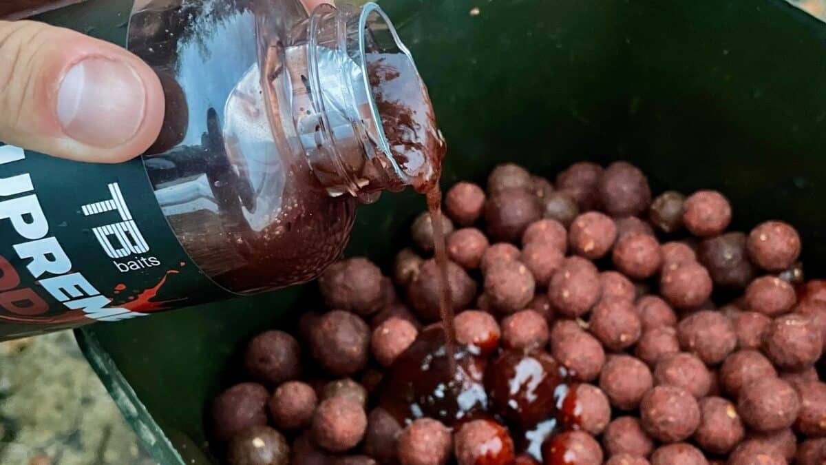 Close-up of a bottle pouring dark liquid into a bucket filled with red-brown fishing bait pellets labeled 'baits'.