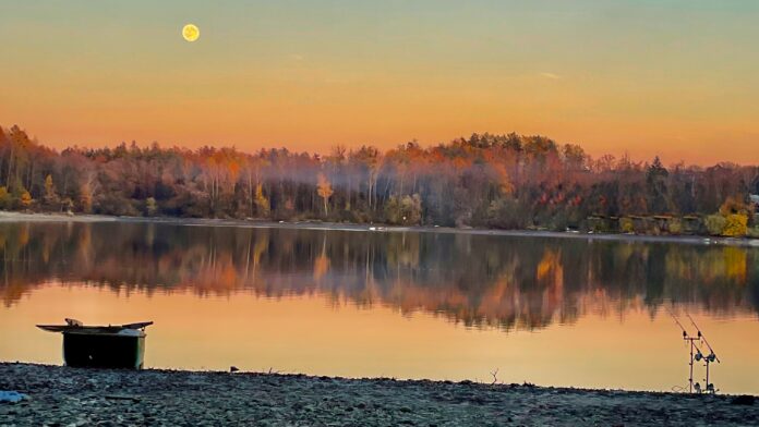 Sunset over a calm lake with a colorful orange-pink sky, a visible full moon, and a tree-lined shore reflecting on the water.