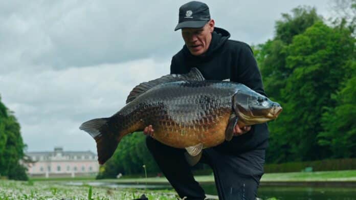 Man wearing a dark hoodie and cap holding a large carp horizontally near a grassy riverbank with trees in the background.
