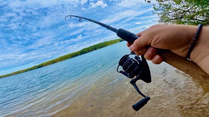 Person fishing from a lakeshore, gripping a spinning rod over calm water with a blue sky overhead.